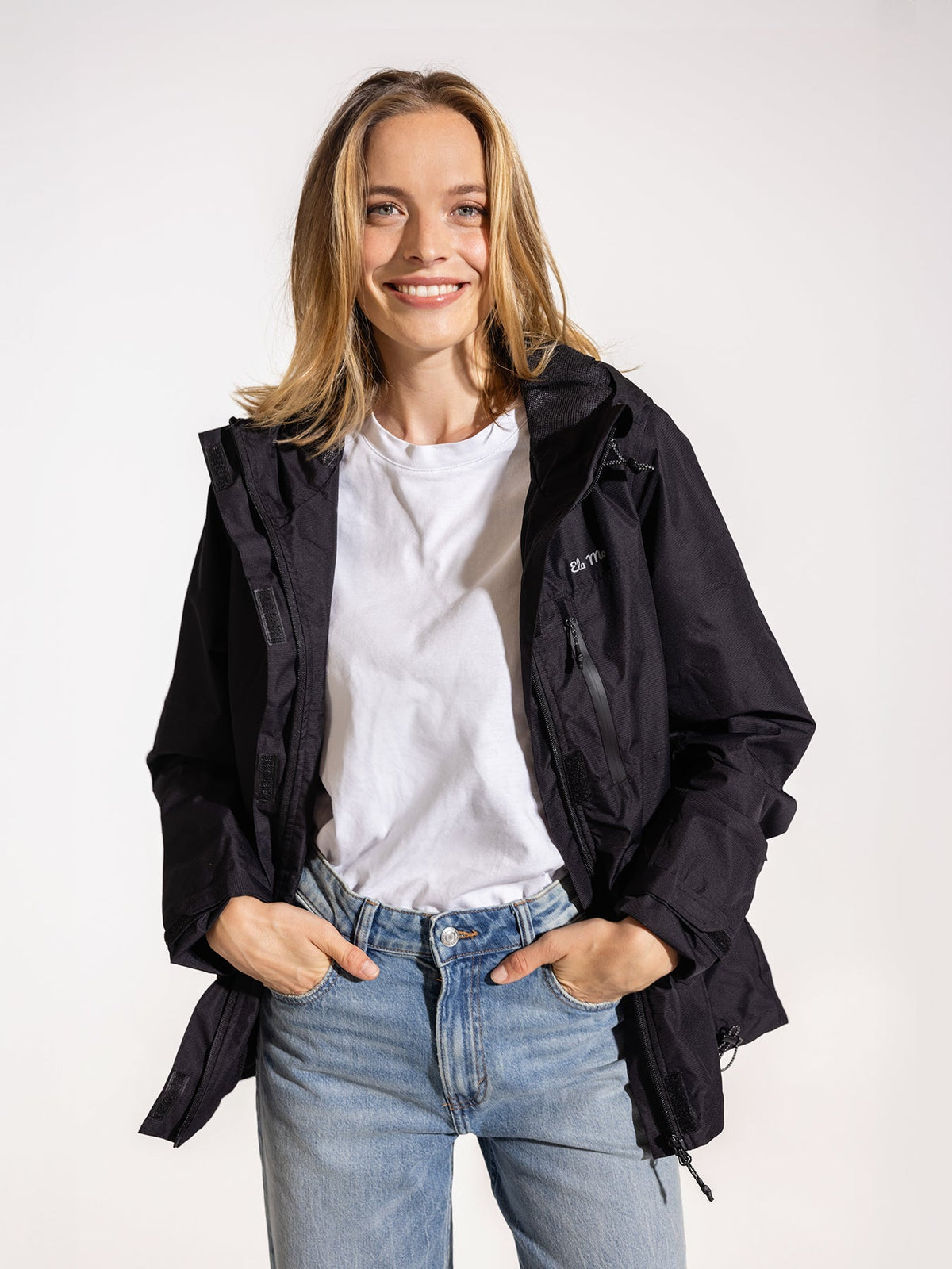 Ela Mo™ Regenjacke | Black. Eine stylische blonde Frau trägt die schwarze Regenjacke, der Reißverschluss ist offen.