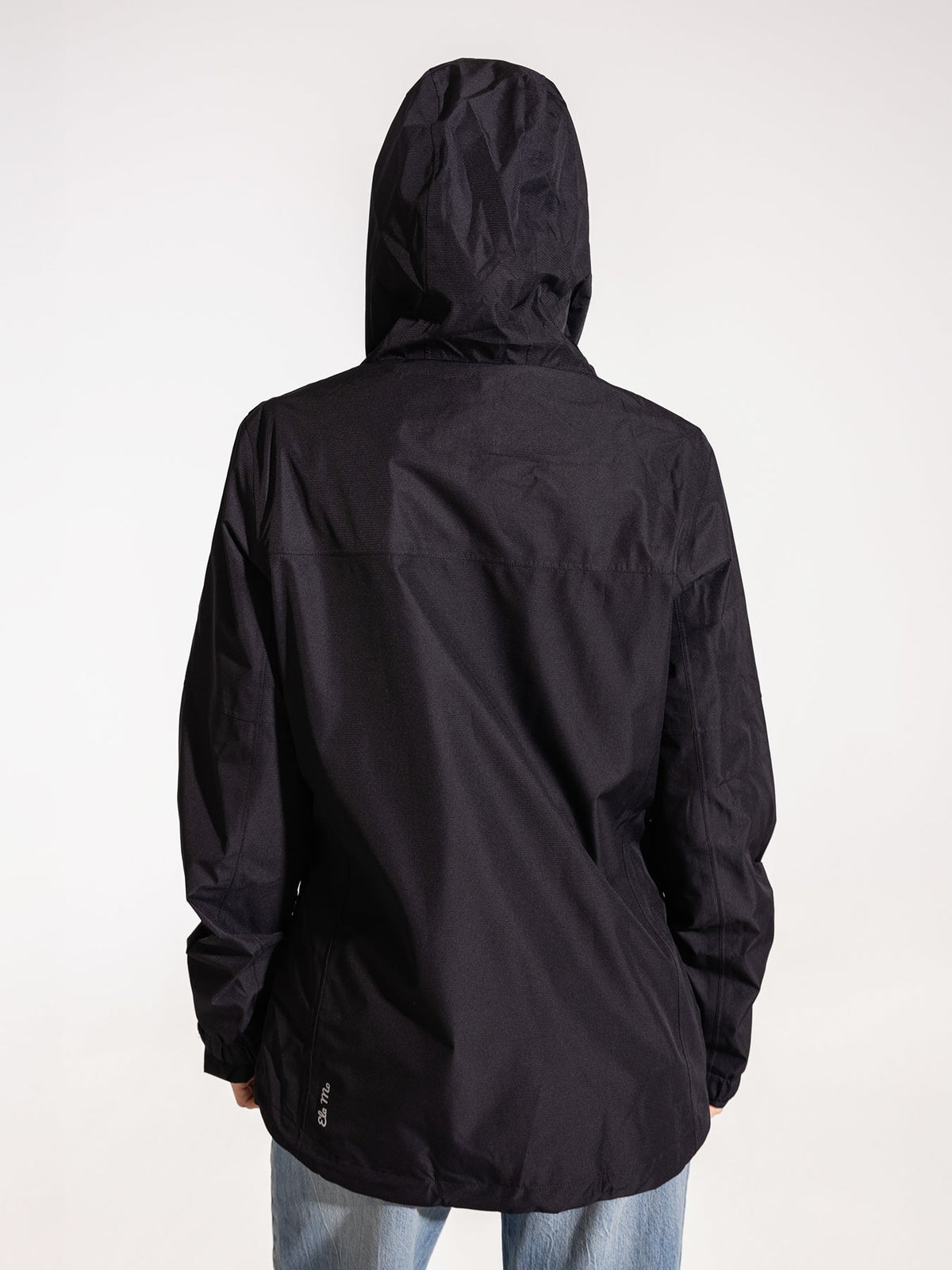 Ela Mo™ Regenjacke | Black. Eine Frau trägt die schwarze Regenjacke, Kapuze auf dem Kopf, Rückenansicht.