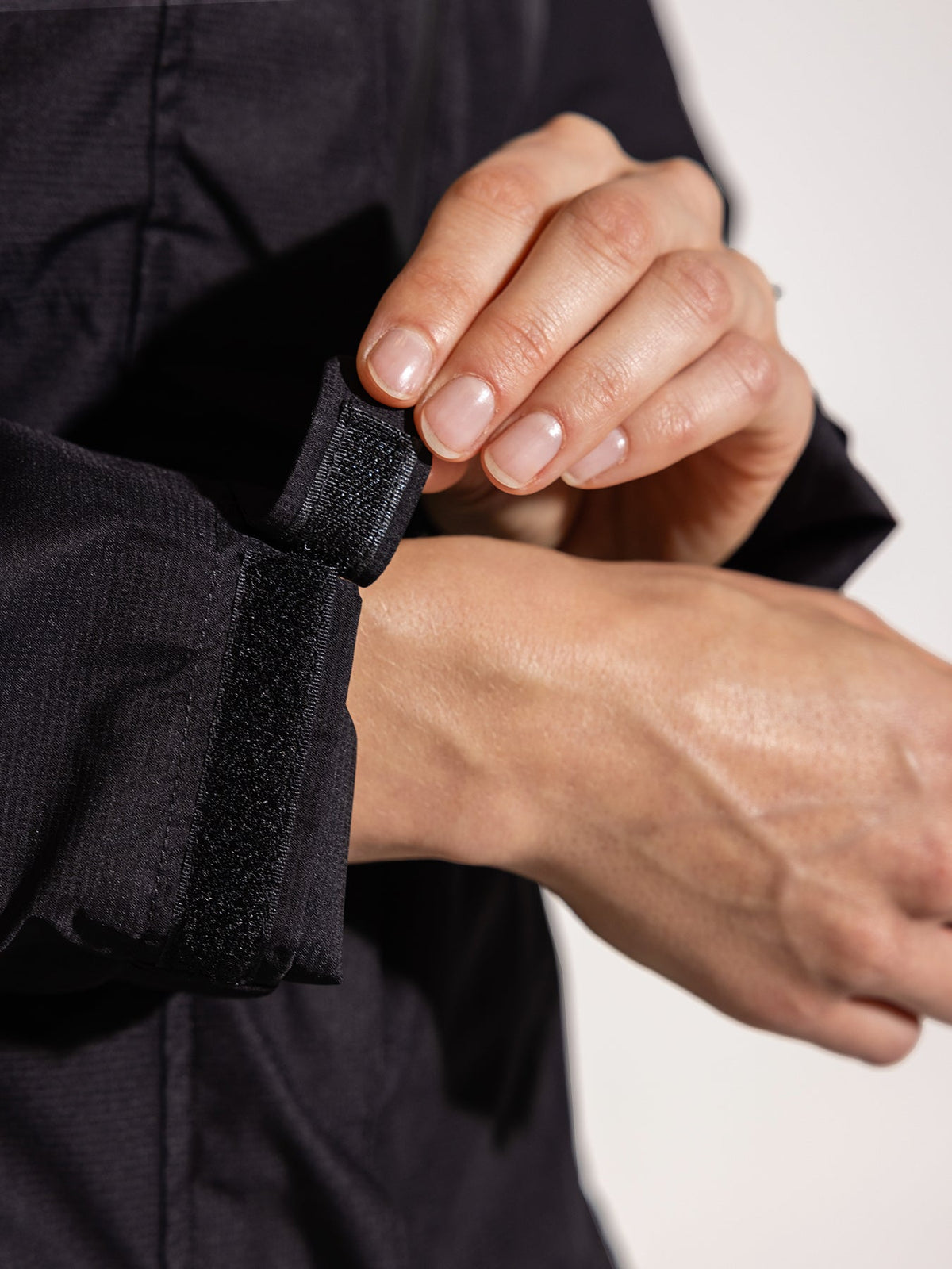 Ela Mo™ Regenjacke | Black. Eine Person präsentiert den verstellbaren Klettverschluss am Ärmel einer schwarzen Regenjacke.