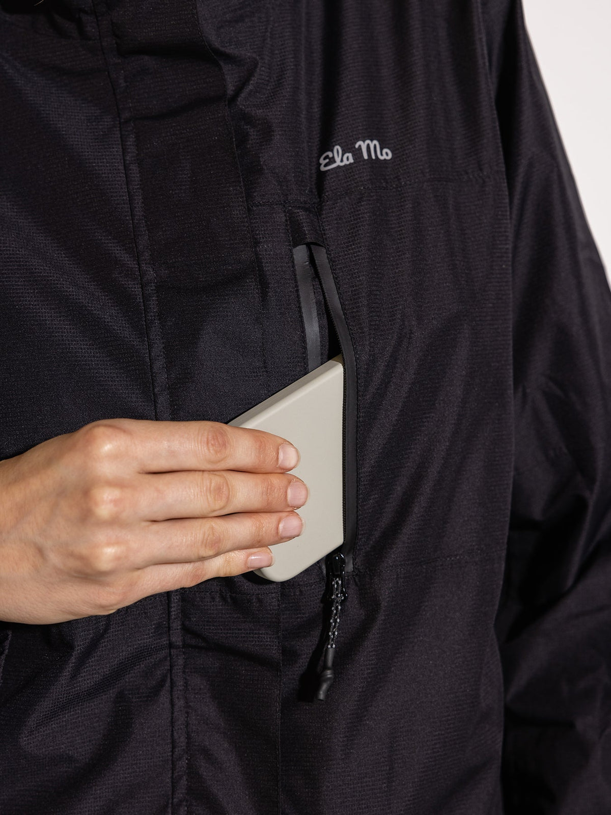 Ela Mo™ Regenjacke | Black. Schwarze Regenjacke mit einer Tasche an der Seite, die mit Reißverschluss geschützt ist.