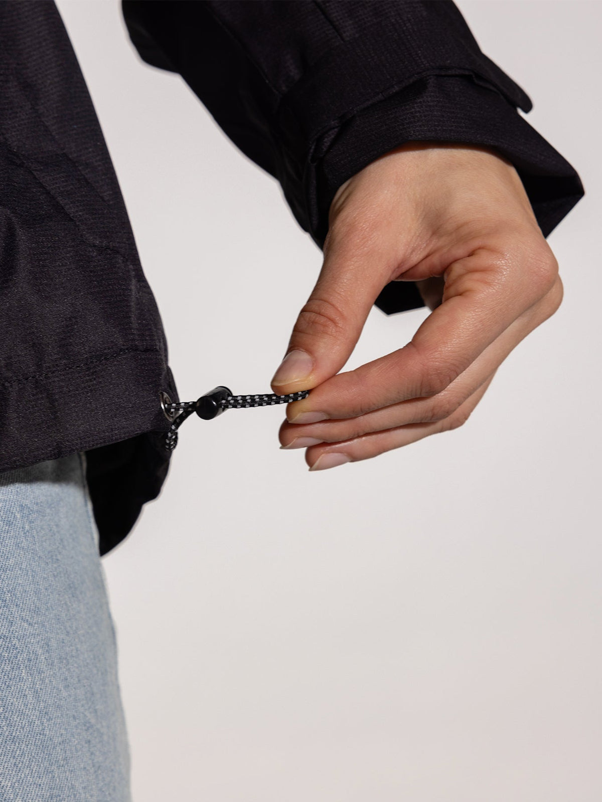 Ela Mo™ Regenjacke | Black. Eine Person mit schwarzer Regenjacke, die den Kordelzug am Saum präsentiert.