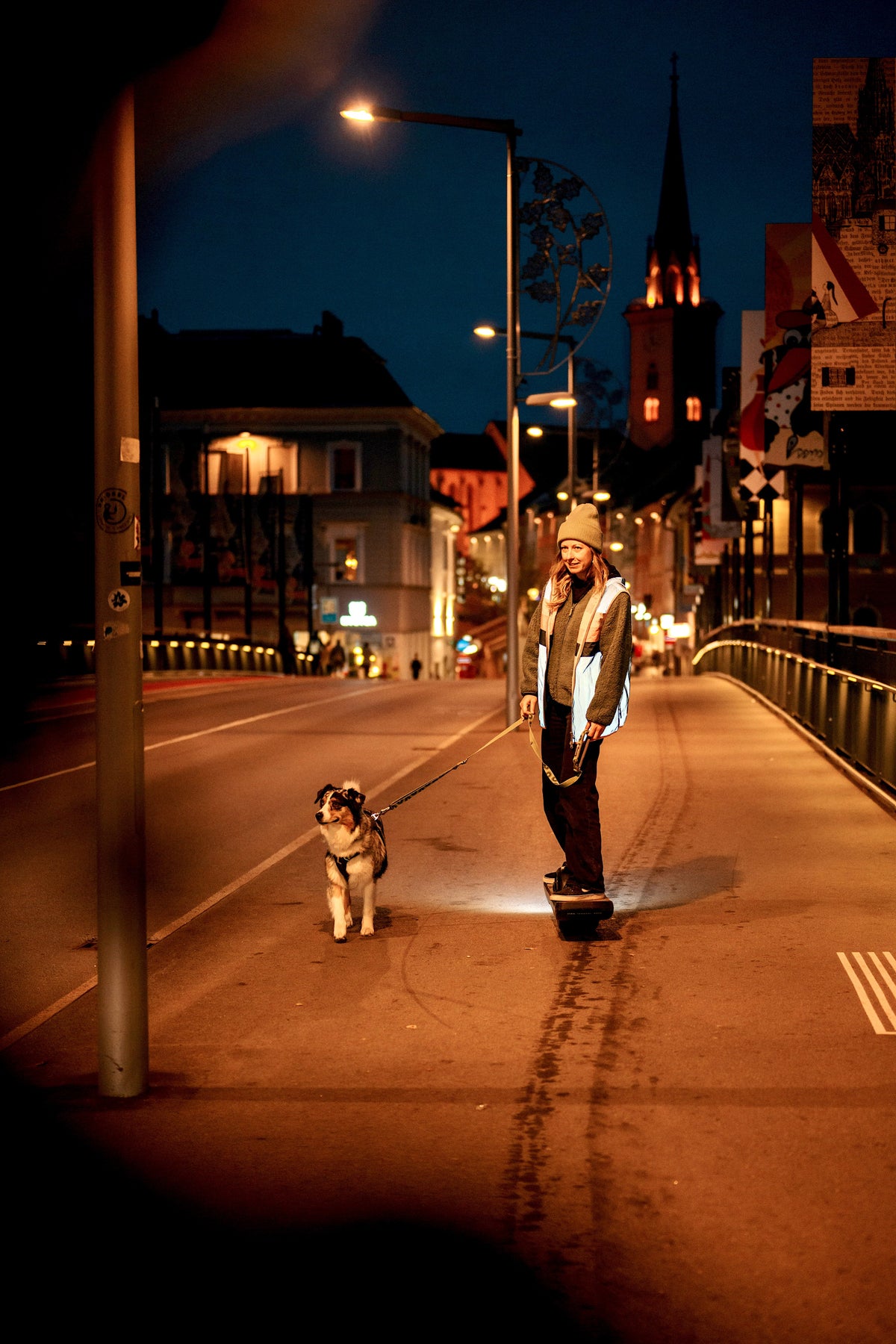 Ela Mo™ Fahrradweste.Eine Frau in reflektierender Fahrradweste in der Nacht mit Hund und Skateboard.