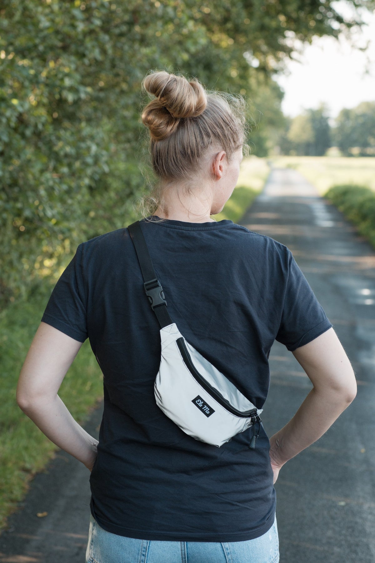 Ela Mo™ Hipbag | Reflective. Eine Frau mit Hipbag an einem sonnigen Tag auf der Wiese, Ansicht von hinten.