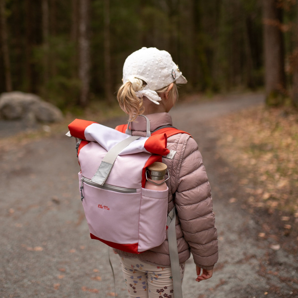 Ela Mo™ Kinderrucksack (6-10 Jahre) | Cotton Candy.Ein Mädchen trägt den rosa Rucksack mit einer Flasche in der Seitentasche