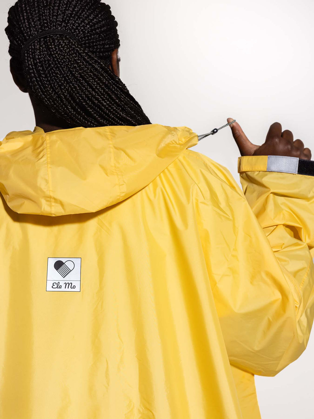 Ela Mo™ Regenponcho | Yellow. Eine Frau im Regenponcho mit Kapuze und Herstellerlogo. Ansicht von hinten