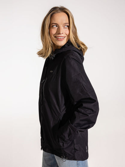 Ela Mo™ Regenjacke | Black. Eine blonde Frau trägt die schwarze Regenjacke mit geschlossenem Reißverschluss, Seitenansicht.