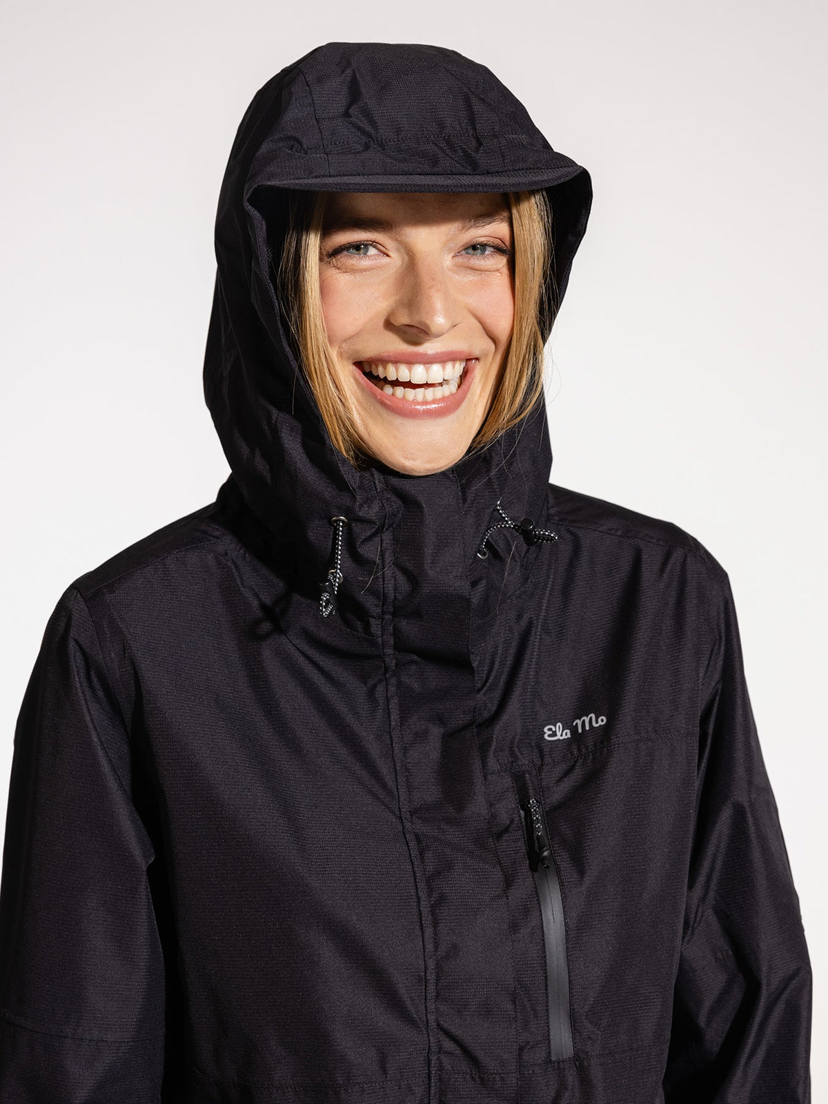 Ela Mo™ Regenjacke | Black. Eine blonde Frau mit schwarzer Regenjacke und Kapuze auf dem Kopf.