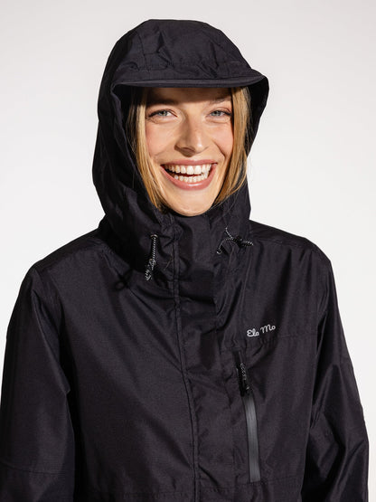 Ela Mo™ Regenjacke | Black. Eine blonde Frau mit schwarzer Regenjacke und Kapuze auf dem Kopf.
