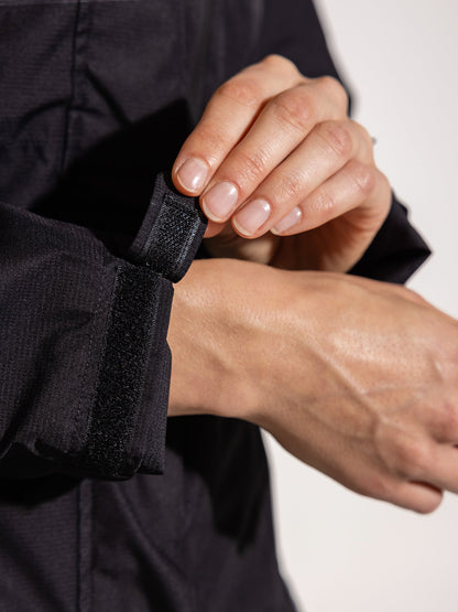 Ela Mo™ Regenjacke | Black. Eine Person präsentiert den verstellbaren Klettverschluss am Ärmel einer schwarzen Regenjacke.