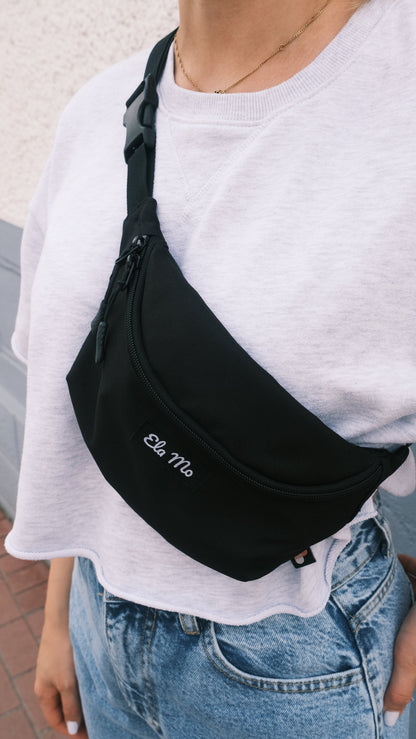 Ela Mo™ Hipbag  | Black. Eine Frau mit Hipbag auf weißem Shirt, alltäglicher Lifestyle.