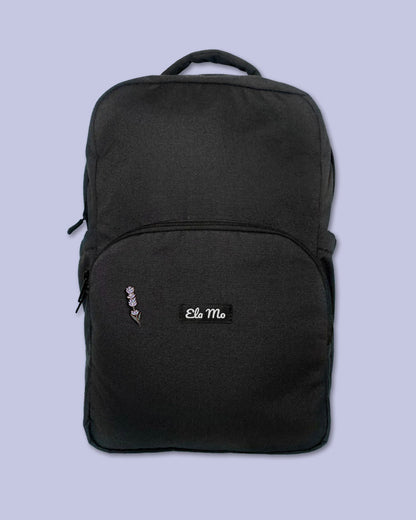 Ela Mo™ Mini Pin für Rucksäcke | Lavender. Ein kleiner Lavendel-Pin am Rucksack, vor lila Hintergrund.