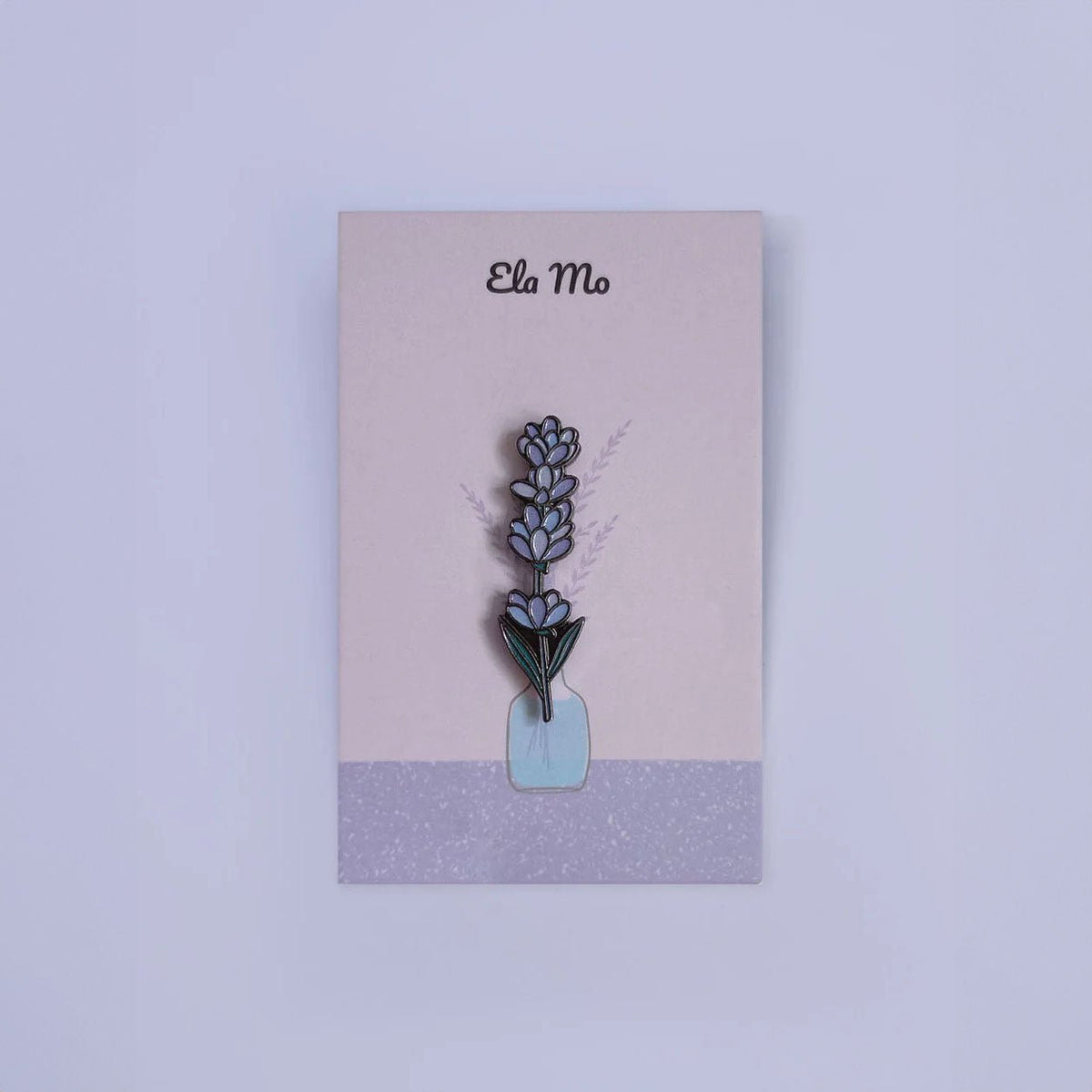 Ela Mo™ Mini Pin für Rucksäcke | Lavender. Ein kleiner Lavendel-Pin auf einer Karte befestigt, vor lila  Hintergrund.