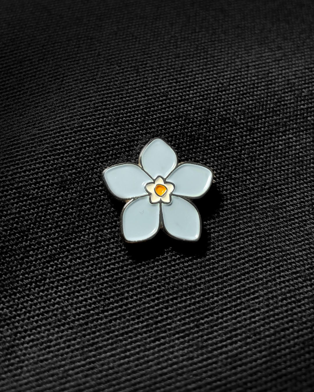Ela Mo™ Mini Pin für Rucksäcke | Vergissmeinnicht. Nahansicht eines kleinen Pins, befestigt auf dem schwarzen Rucksack.