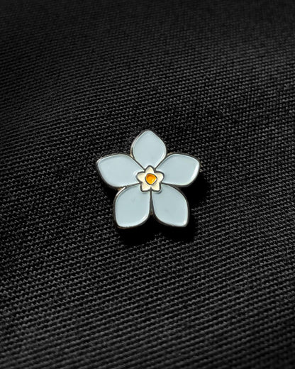 Ela Mo™ Mini Pin für Rucksäcke | Vergissmeinnicht. Nahansicht eines kleinen Pins, befestigt auf dem schwarzen Rucksack.