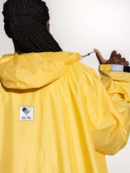Ela Mo™ Regenponcho | Yellow. Eine Frau im Regenponcho mit Kapuze und Herstellerlogo. Ansicht von hinten