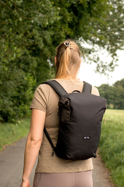 Ela Mo™ Rolltop Lite | Black. Eine Frau mit dem Rucksack auf der Wiese, Ansicht von hinten.